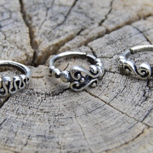Peut inclure: Trois anneaux de nez en argent avec des motifs ornés différents. Les anneaux sont sur une surface en bois.
