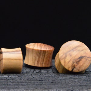 May include: Three wooden ear plugs with a natural wood grain pattern. The plugs vary in shape, with one cylindrical, one hourglass, and one flat-faced. The wood tones range from light tan to medium brown, set against a dark background.