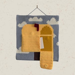 Pode incluir: Uma tapeçaria quadrada de crochê apresentando uma caixa de correio amarela com uma bandeira vermelha contra um céu azul claro com nuvens brancas. A caixa de correio tem um bolso funcional contendo um cartão retangular branco e uma aba.