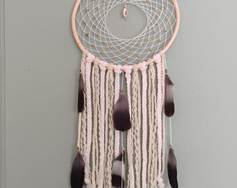 Puede incluir: Un atrapasueños artesanal con un marco circular de madera y un diseño de red tejida. El atrapasueños presenta hilo blanco, cuentas de madera y plumas colgantes en tonos negros y grises. Está diseñado para la decoración de paredes.