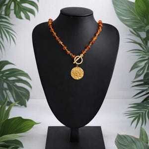 Può includere: Una collana dorata con perline color ambra e un ciondolo strutturato. La collana è esposta su un manichino nero su uno sfondo di fogliame verde. Il ciondolo è rotondo con una superficie dettagliata e una chiusura dorata.