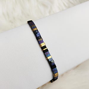 Peut inclure: Un bracelet de perles composé de petites perles rectangulaires bleues, violettes, noires et dorées. Le bracelet est présenté sur une surface blanche, mettant en valeur son design géométrique et sa palette de couleurs. Le fermoir du bracelet est doré.