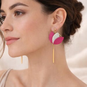 Può includere: Primo piano di una donna che indossa orecchini a forma di fiore rosa e argento con dettagli color oro. Gli orecchini hanno una lunga barra dorata che pende dal fiore.