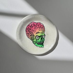 May include: A round, off-white ceramic trinket dish featuring a central illustration of a stylized skull with an oversized, brain-like cranium. The skull is colored with a vibrant gradient transitioning from pink at the top to green at the base.