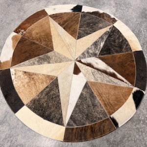 May include: A circular rug constructed from stitched cowhide patches in a geometric star pattern. The hide features a natural color palette of white, cream, tan, brown, and black, with varying textures and hair lengths across the individual segments.