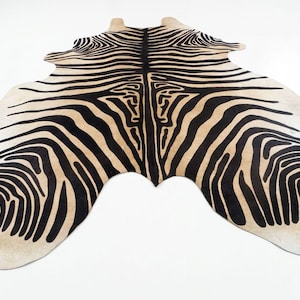 May include: A zebra-print rug with a beige background and bold black stripes. The rug has an irregular shape, mimicking the form of an animal hide. The design features a symmetrical pattern, creating a striking visual effect. This decorative rug is suitable for home decor.