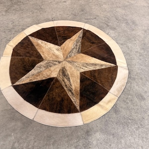 May include: A circular rug constructed from stitched cowhide panels in shades of dark brown, tan, and cream. The design features a central five-pointed star pattern with alternating dark and light sections, finished with a solid cream-colored border.