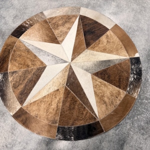 May include: A circular rug constructed from stitched panels of cowhide in varying shades of brown, tan, cream, and black. The panels are arranged in a geometric star pattern that radiates from the center toward the outer circular edge.