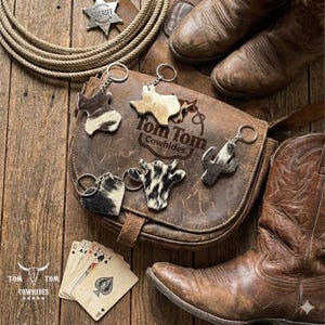 May include: Rustic western-themed image featuring leather keychains, a leather bag, cowboy boots, and playing cards. Keychains include cowhide and cactus designs. The bag is branded "Tom Tom Cowhides". A sheriff's badge and rope complete the scene.