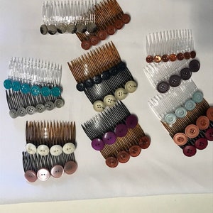 May include: Ten hair combs with different coloured buttons attached. The combs are made of plastic and have a variety of colours, including black, white, brown, blue, and purple.