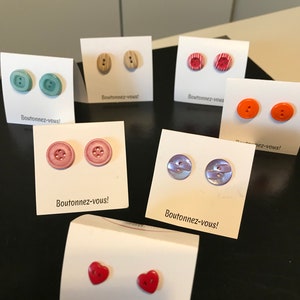 Puede incluir: Surtido de pendientes de botón coloridos mostrados en tarjetas blancas con el texto "Boutonnez-vous!" que se traduce como "¡Abotónate!" en inglés. Los pendientes están hechos de varios materiales, incluyendo madera, plástico y metal.