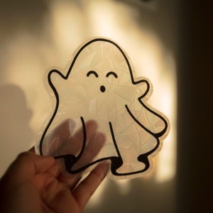 May include: A white iridescent ghost sticker with black outline and a smiling face. The ghost has two small arms and two small legs.