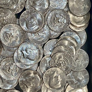 May include: A close-up shot of a collection of silver Kennedy half-dollar coins. The coins are scattered, showing the profile of John F. Kennedy and the American eagle. The coins are in a dark container.