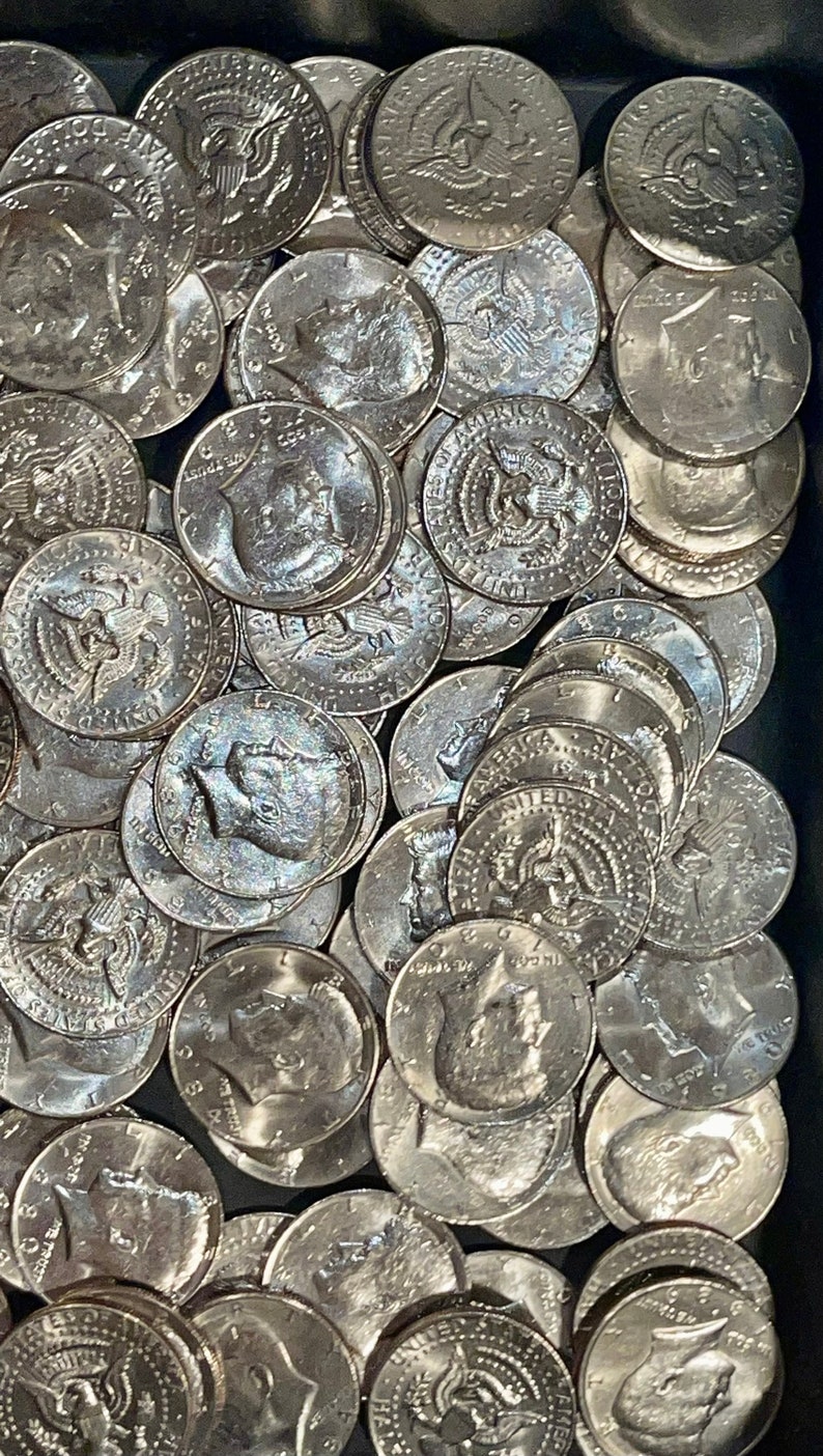 May include: A close-up shot of a collection of silver Kennedy half-dollar coins. The coins are scattered, showing the profile of John F. Kennedy and the American eagle. The coins are in a dark container.