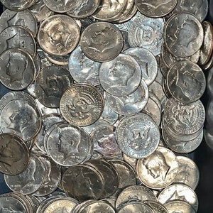 May include: A close-up shot of a pile of silver Kennedy half-dollar coins. The coins are scattered, showing the profile of John F. Kennedy and the words "IN GOD WE TRUST" and the year 1972 or 1971.