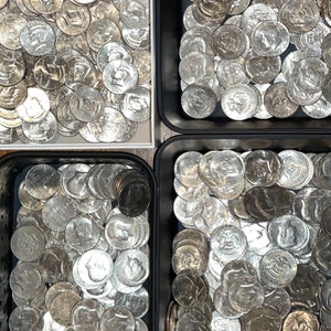 May include: An overhead shot shows four containers filled with silver half-dollar coins. The coins are arranged in a seemingly random pattern, with the profiles of historical figures visible on their surfaces. The containers are black and white.