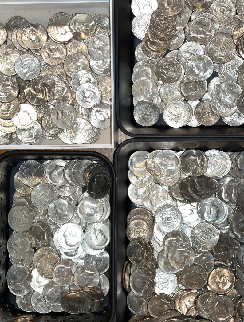 May include: An overhead shot shows four containers filled with silver half-dollar coins. The coins are arranged in a seemingly random pattern, with the profiles of historical figures visible on their surfaces. The containers are black and white.