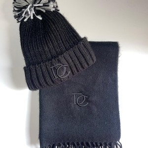 May include: A black knitted beanie hat with a pom-pom and a matching scarf. The beanie has a ribbed brim and a logo. The scarf is rectangular with a logo and fringe at the bottom. The pom-pom is white, black, and grey.