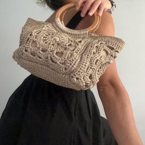 Pu&ograve; includere: Una borsa a mano in crochet beige con manico in legno. La borsa ha un design delicato e traforato.