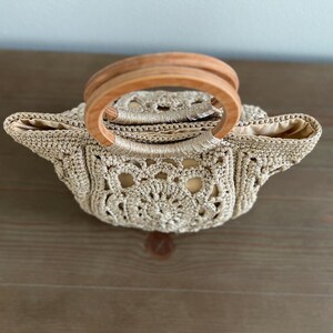 Pu&ograve; includere: Una borsa all'uncinetto con un manico in legno. La borsa &egrave; beige e ha un delicato motivo a pizzo.
