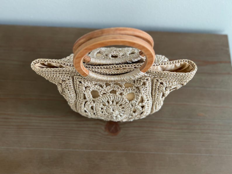 Pu&ograve; includere: Una borsa all'uncinetto con un manico in legno. La borsa &egrave; beige e ha un delicato motivo a pizzo.