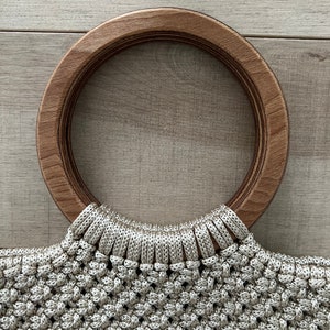 Puede incluir: Un bolso de mano de crochet beige con un asa redonda de madera grande.