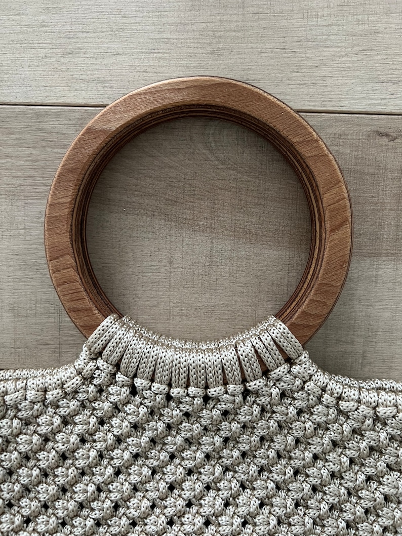 Puede incluir: Un bolso de mano de crochet beige con un asa redonda de madera grande.