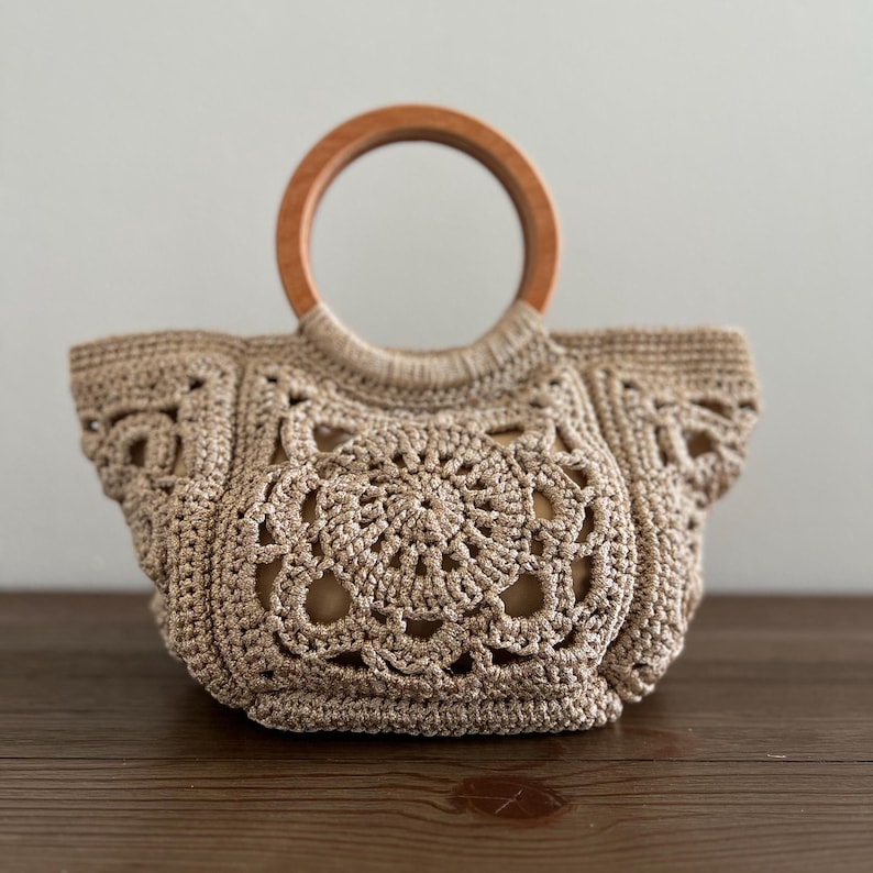 Pu&ograve; includere: Una borsa a mano in crochet beige con un manico in legno. La borsa ha un grande disegno circolare al centro.
