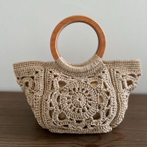 Pu&ograve; includere: Una borsa a mano in crochet beige con manico in legno. La borsa presenta un motivo circolare al centro e un bordo a festoni.