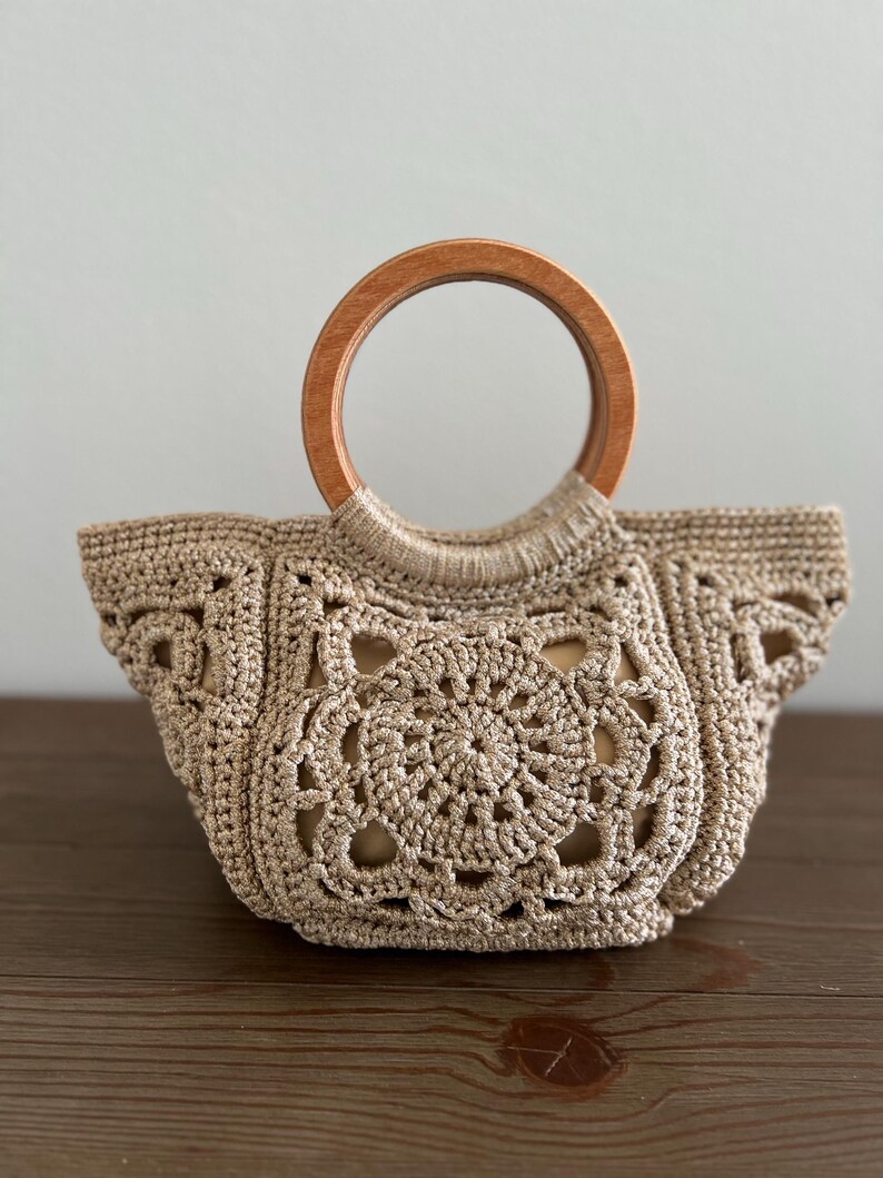 Pu&ograve; includere: Una borsa a mano in crochet beige con manico in legno. La borsa presenta un motivo circolare al centro e un bordo a festoni.