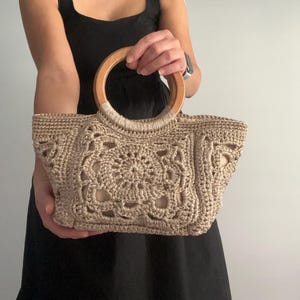 Pu&ograve; includere: Una borsa a mano in crochet beige con manico in legno. La borsa ha una forma quadrata e un design delicato e intricato.