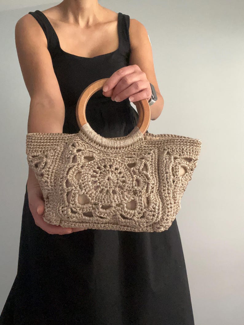 Pu&ograve; includere: Una borsa a mano in crochet beige con manico in legno. La borsa ha una forma quadrata e un design delicato e intricato.