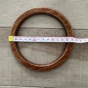 Puede incluir: Un aro de bordado de madera marr&oacute;n con un di&aacute;metro de 35,5 cm. El aro est&aacute; sobre una superficie de madera marr&oacute;n claro.