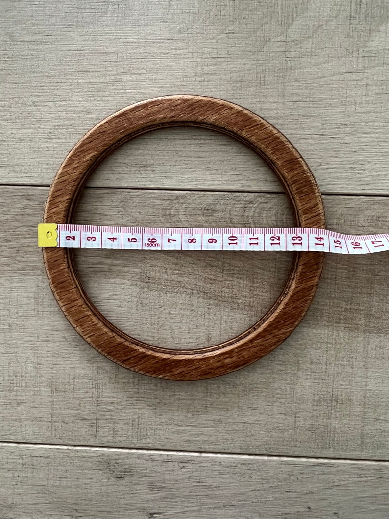 Puede incluir: Un aro de bordado de madera marr&oacute;n con un di&aacute;metro de 35,5 cm. El aro est&aacute; sobre una superficie de madera marr&oacute;n claro.