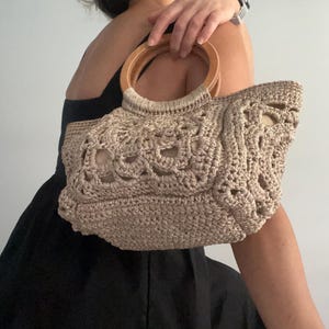 Pu&ograve; includere: Una borsa a mano in crochet beige con un manico in legno. La borsa ha un design delicato e traforato.