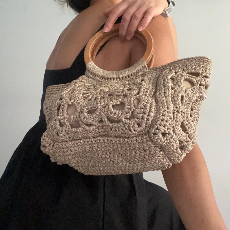 Pu&ograve; includere: Una borsa a mano in crochet beige con un manico in legno. La borsa ha un design delicato e traforato.