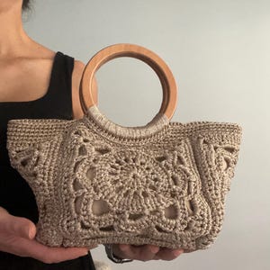 Pu&ograve; includere: Una borsa a mano in crochet beige con manico in legno. La borsa ha un design delicato e traforato.