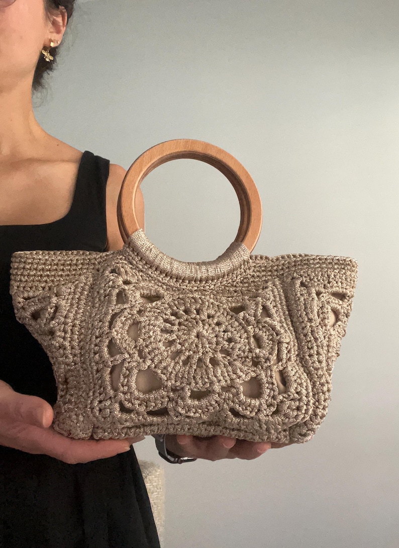 Pu&ograve; includere: Una borsa a mano in crochet beige con manico in legno. La borsa ha un design delicato e traforato.