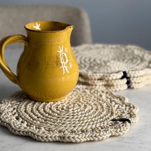 Könnte beinhalten: Ein Set aus vier gehäkelten runden Untersetzern in Beige mit schwarzem Besatz. Ein gelber Keramik-Krug steht auf einem der Untersetzer.