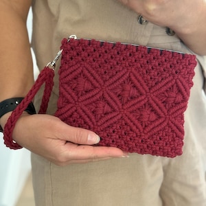 Pode incluir: Uma clutch de macramé bordô com um padrão geométrico de diamantes. A clutch tem uma alça de pulso combinando e um fecho de zíper prateado. Uma pessoa com um macacão bege a segura.