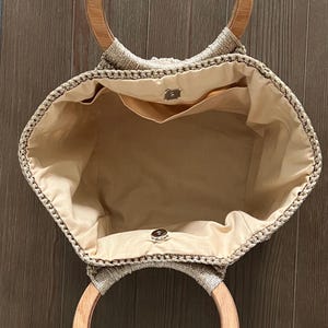 Pu&ograve; includere: Una borsa tote in crochet beige con manici in legno. La borsa ha una fodera in tessuto beige e una piccola tasca interna.