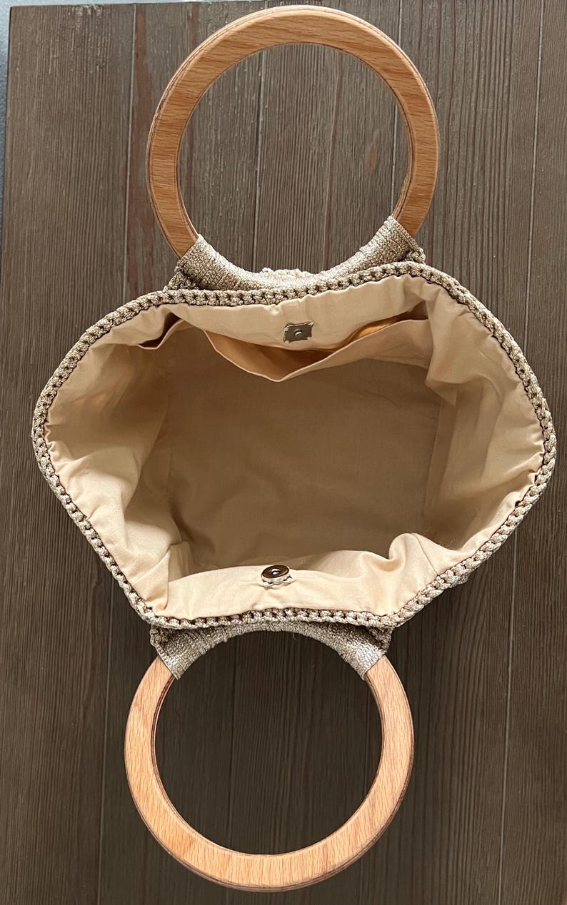 Pu&ograve; includere: Una borsa tote in crochet beige con manici in legno. La borsa ha una fodera in tessuto beige e una piccola tasca interna.