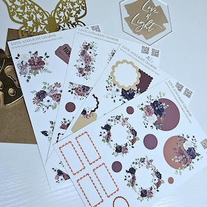 Peut inclure: Plusieurs autocollants floraux avec des fleurs bordeaux, blanches et bleu marine. Les autocollants ont différentes formes, notamment des cercles, des couronnes et des cadres. Un papillon doré et une enseigne "Love & Light" sont également présents.