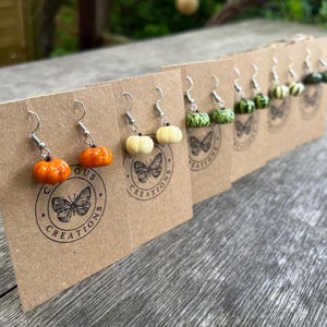 May include: A selection of pumpkin earrings in shades of orange, white, and green, displayed on brown cards. Each card features a butterfly logo and the text "CURIOUS CREATIONS". The earrings have silver-coloured hooks. The earrings are a seasonal autumn theme.
