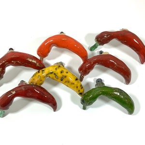 May include: A collection of six ceramic pepper-shaped door handles in various colors, including red, green, yellow, and orange. The handles are all in the shape of a pepper and have a slightly curved shape.