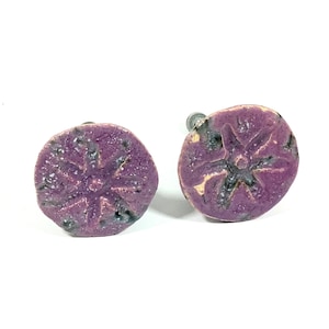 Op de afbeelding: Twee paarse keramische ladeknopjes met een stervormig ontwerp. De knoppen hebben een rustieke, handgemaakte uitstraling.