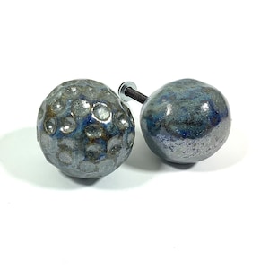 Op de afbeelding: Twee blauwe en grijze keramische knoppen met een zilveren metalen schroef. De knoppen hebben een getextureerd oppervlak.