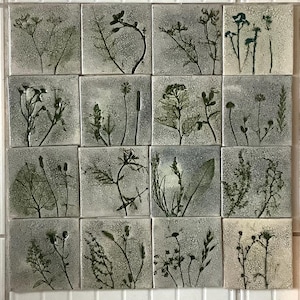 Op de afbeelding: Een verzameling van zestien vierkante keramische tegels, elk met een uniek geperst botanisch ontwerp. De planten zijn in tinten groen en bruin tegen een gevlekte grijze achtergrond. De tegels zijn in een rasterpatroon gerangschikt.