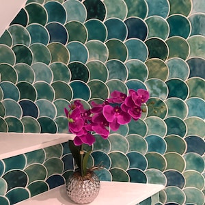 May include: A silver vase with a textured surface holds a bouquet of pink orchid flowers. The vase is sitting on a white shelf in front of a wall covered in green and blue fish scale tiles.