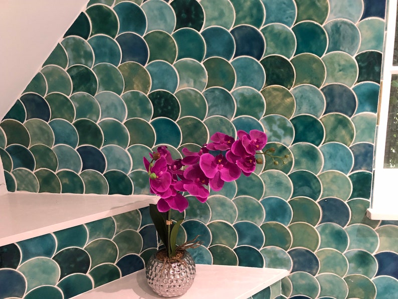 May include: A silver vase with a textured surface holds a bouquet of pink orchid flowers. The vase is sitting on a white shelf in front of a wall covered in green and blue fish scale tiles.